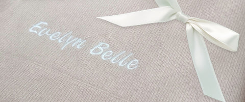 Close-up of a pink cashmere baby blanket with the words 'Evelyn Belle' embroidered in white script, tied with a white satin ribbon bow.