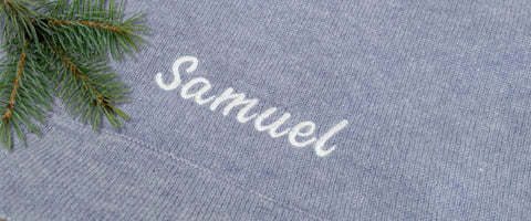 A close-up of a blue cashmere baby blanket with the name 'Samuel' embroidered in white script, styled with a pine branch in the top-left corner.