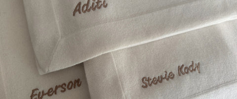 Close-up of folded cream cashmere baby blankets with embroidered names in bronze thread: 'Aditi', 'Everson', and 'Stevie Kody'.