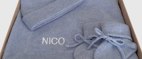 A light blue cashmere baby set, including a blanket, hat and booties, neatly folded inside a cardboard gift box. The blanket features the name 'NICO' embroidered in white on the front.